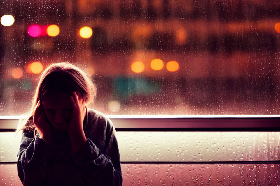 Person in distress by rainy window with soft bokeh lights.