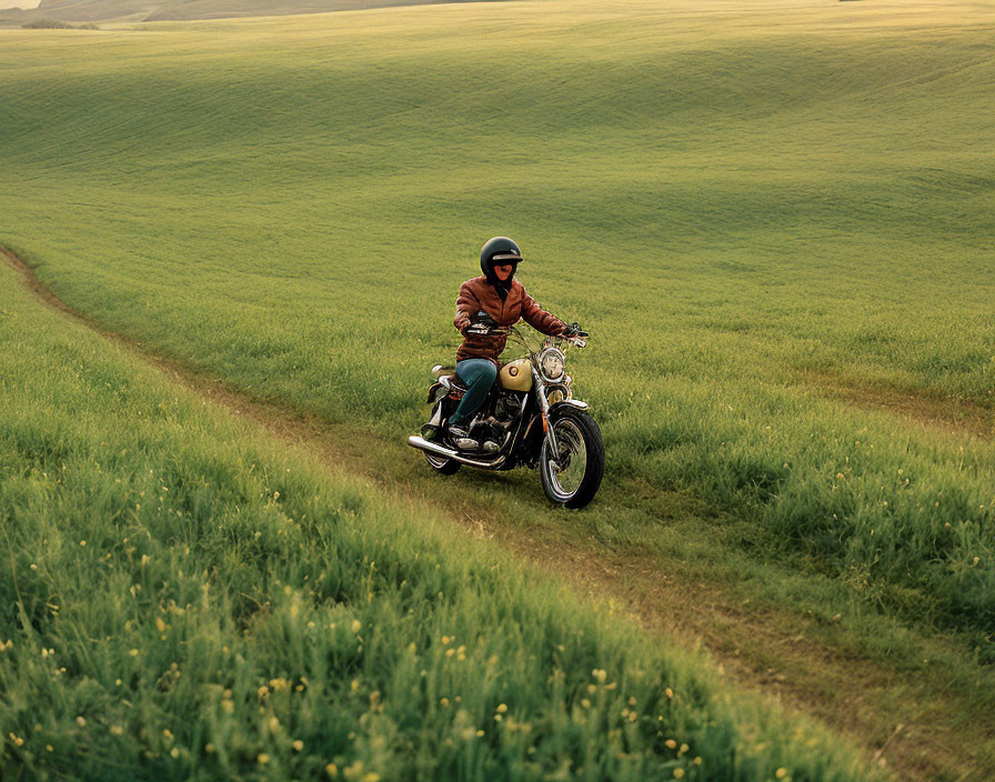 Person riding classic motorcycle on curving dirt path through lush green field