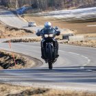 Person in black outfit riding motorcycle on icy road with stationary bike nearby