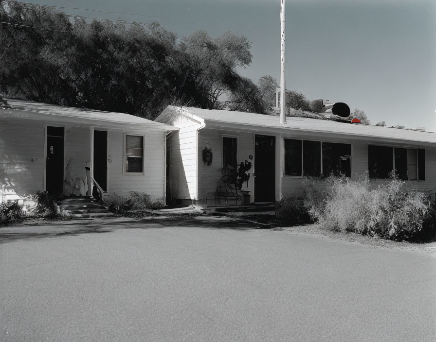 Monochrome image of single-story house with flagpole, bushes, trees & clear sky