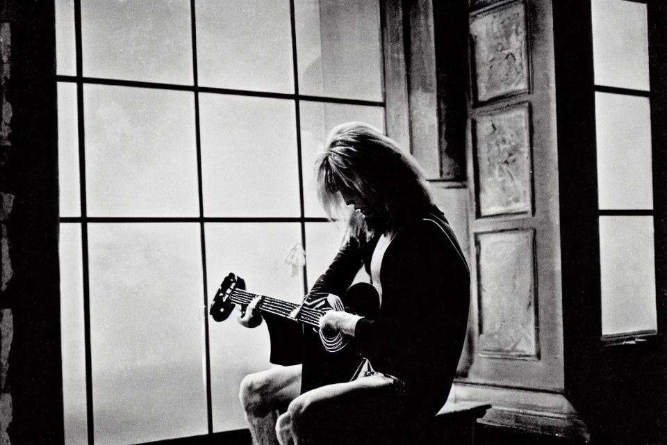 Silhouetted figure playing guitar by large window in dimly lit room