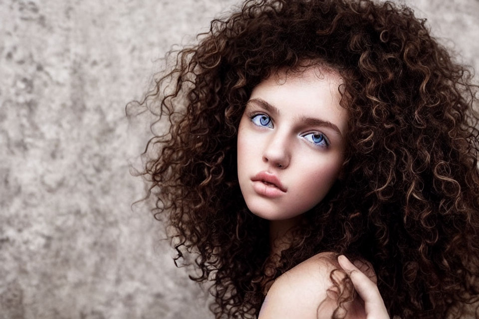 Young woman with voluminous curly hair and striking blue eyes against neutral textured backdrop