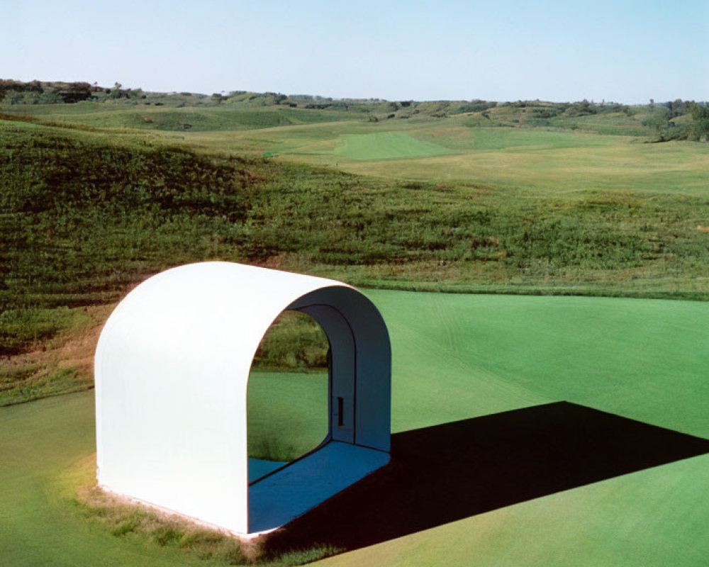 Small white arch-shaped building in vast green landscape under clear blue sky