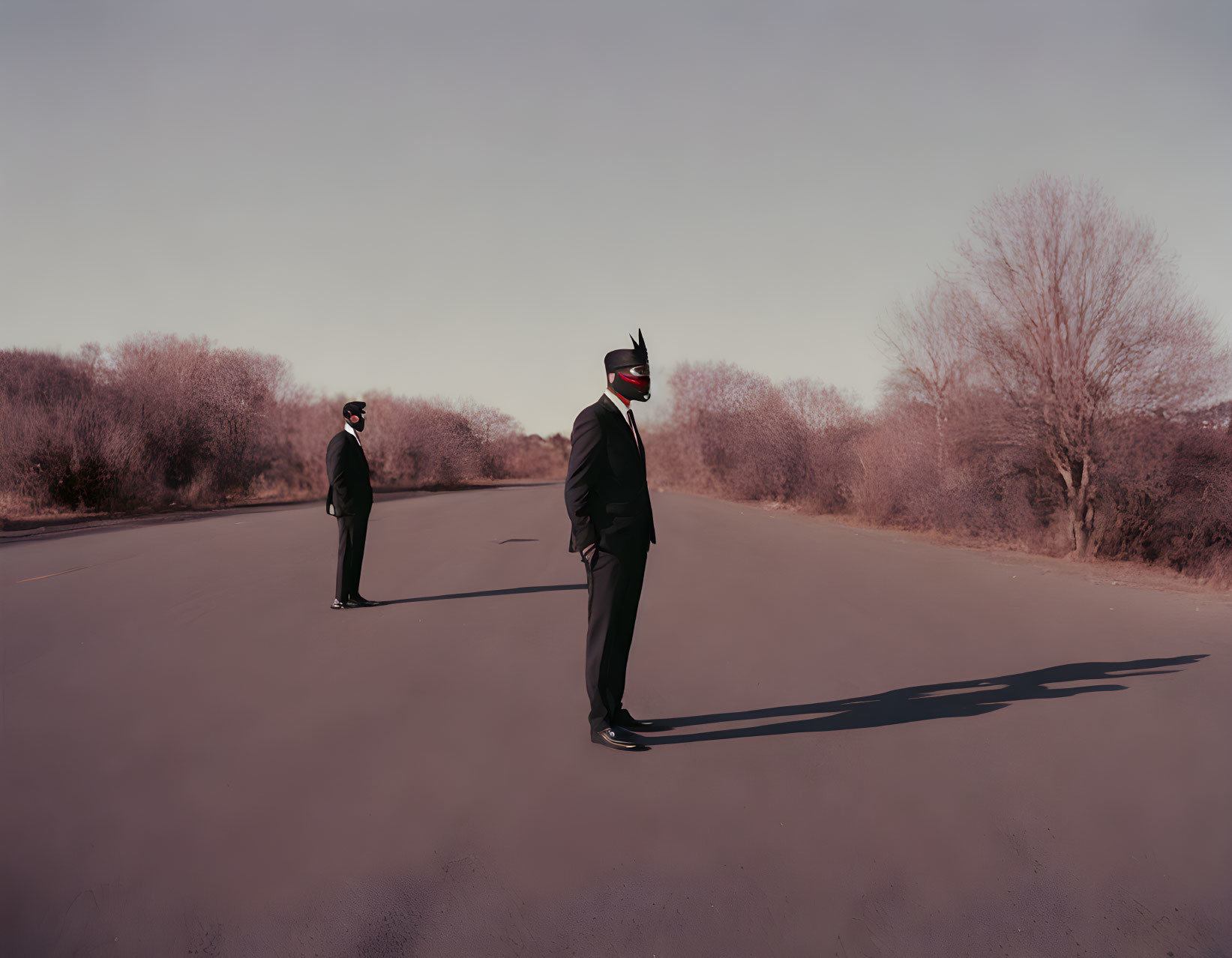 Two individuals in bird masks and suits on deserted road with bare trees