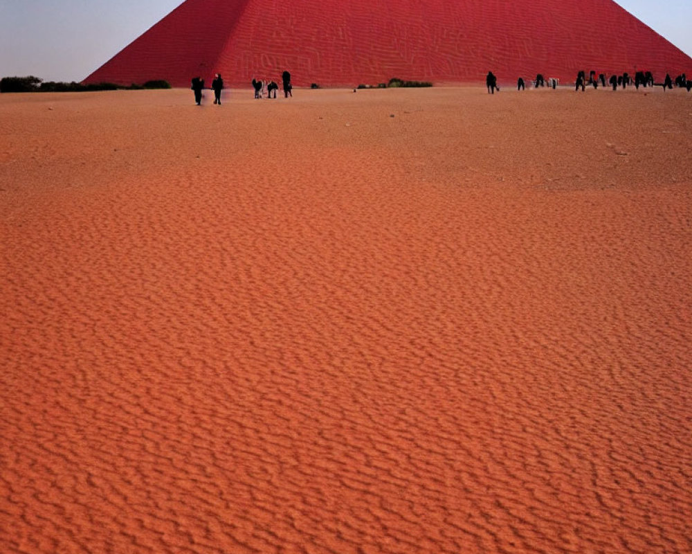 People walking near a large red sand dune under a clear blue sky