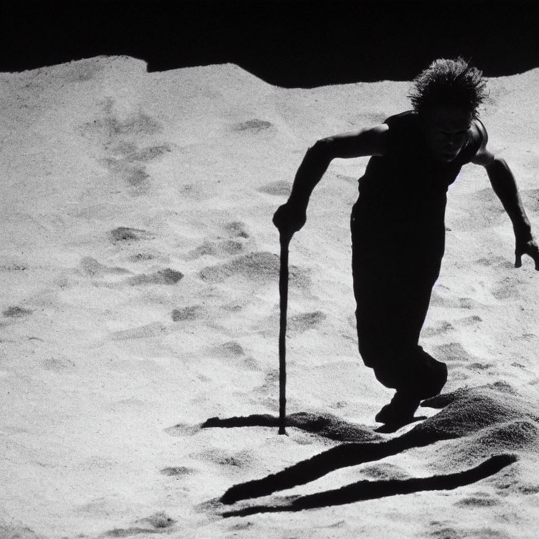 Spiky-haired person walking on sand with long shadow and stick