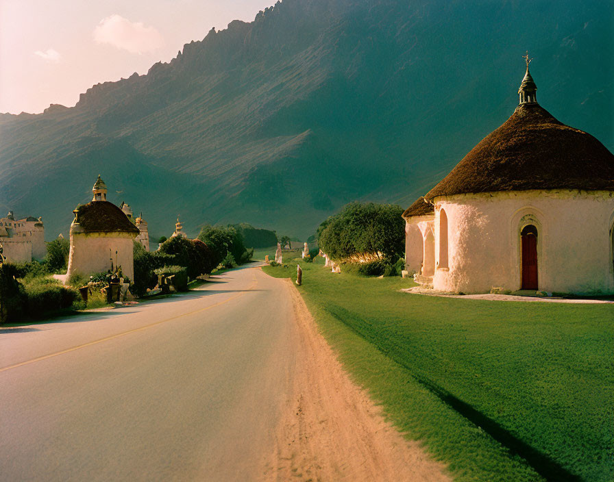 Serene road with white buildings, chapels, green grass, mountains, soft-lit sky