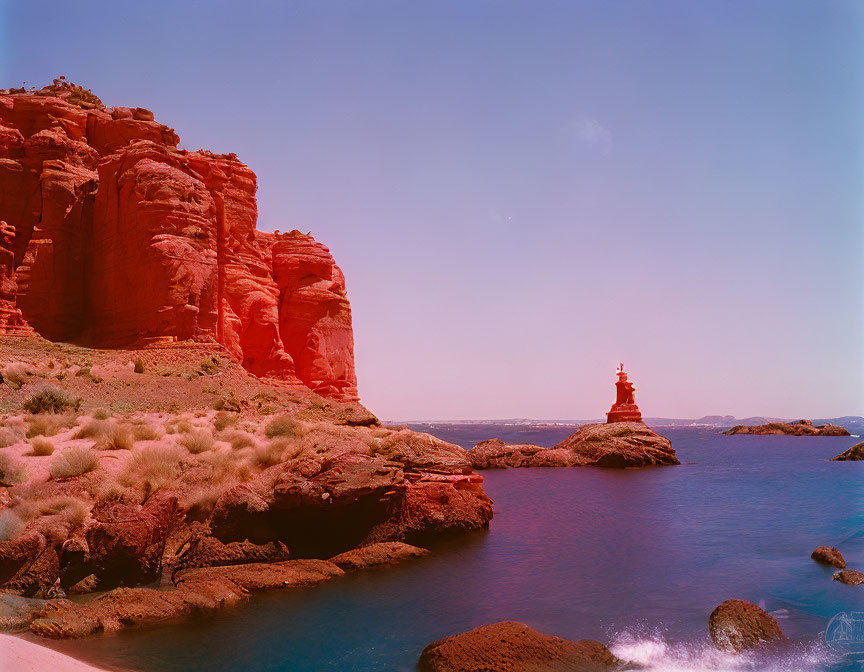 Red rock formation by calm blue water under clear sky with green shrubs