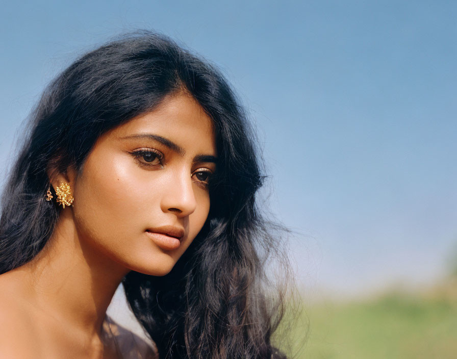 Dark-haired woman in golden earrings gazing into the distance under a blue sky