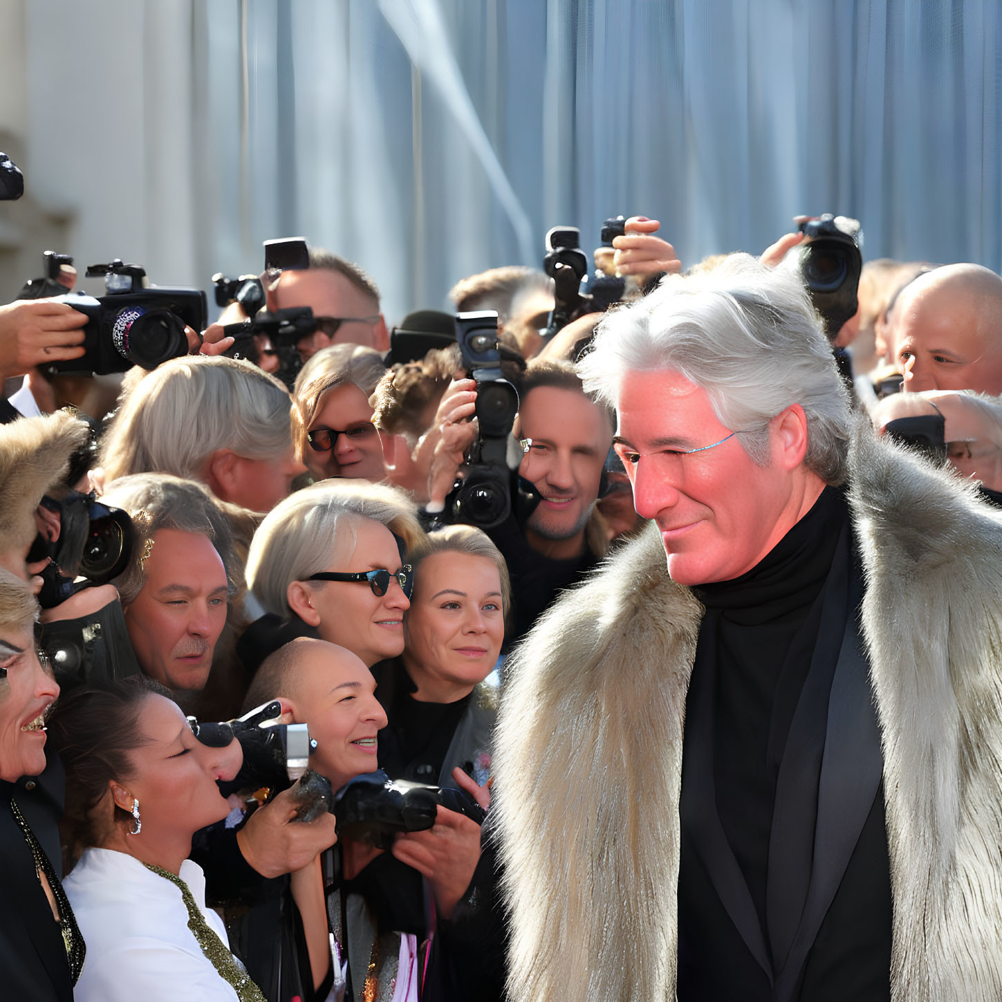 Silver-haired man in fur coat and turtleneck smiles amid photographers.