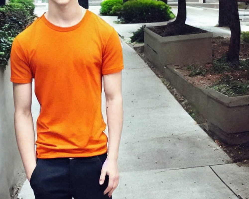 Young man with dark hair in orange t-shirt and black pants on sidewalk with trees and building.