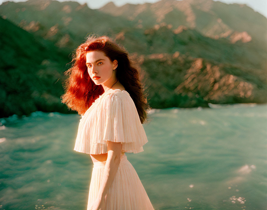 Red-haired woman in pale dress by the sea and rocky hills under warm sunlight