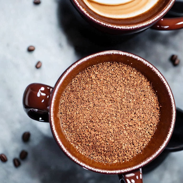 Two mugs with frothy coffee and coffee grounds on textured surface