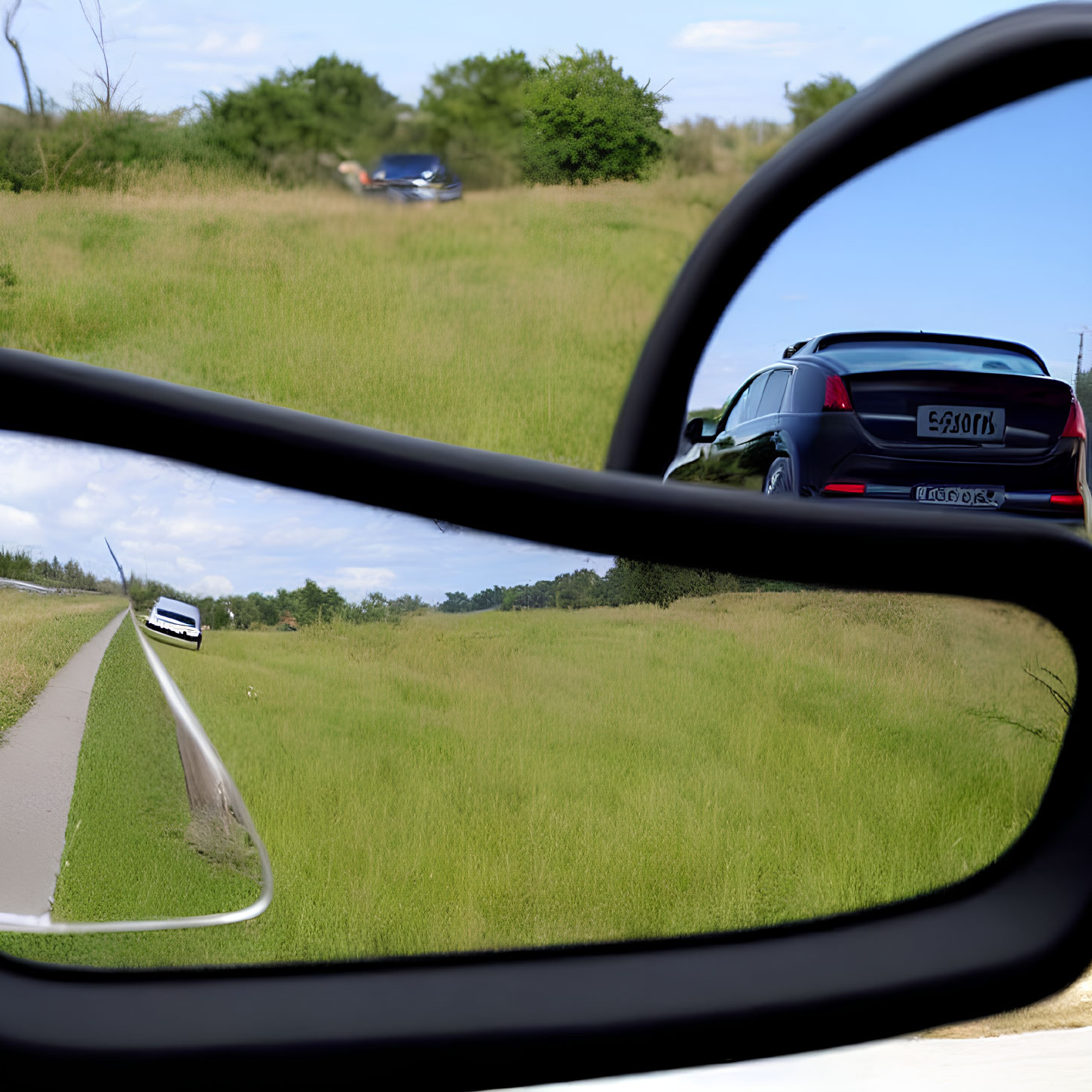 Road and car reflected in side-view mirror against grassy field and sky