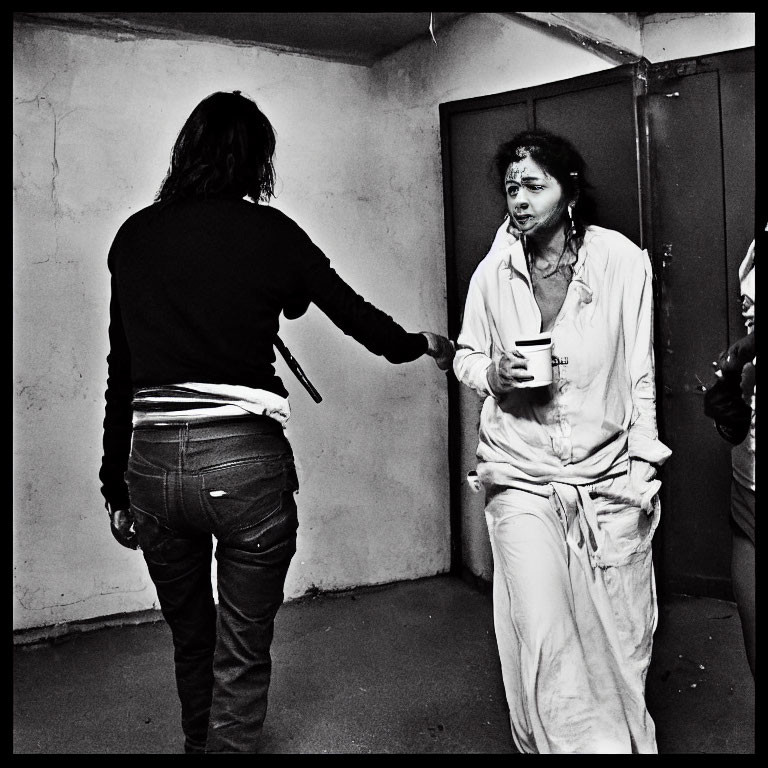 Distressed person in white outfit receives cup from person in black top in bleak room