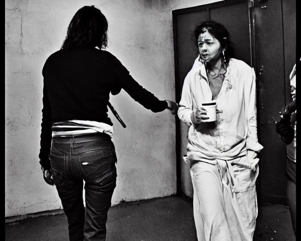 Distressed person in white outfit receives cup from person in black top in bleak room