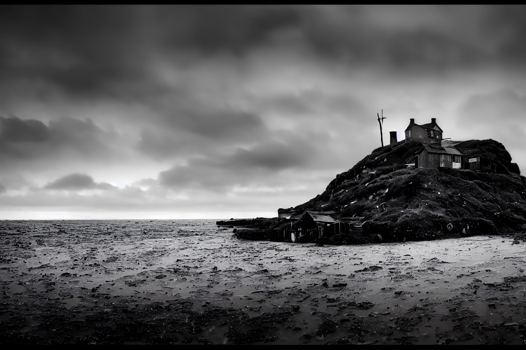 Monochrome image: desolate house on rocky outcrop by the sea under dramatic sky