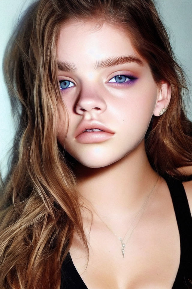 Young woman with wavy brown hair and blue eyes wearing black top and delicate necklace gazes at camera