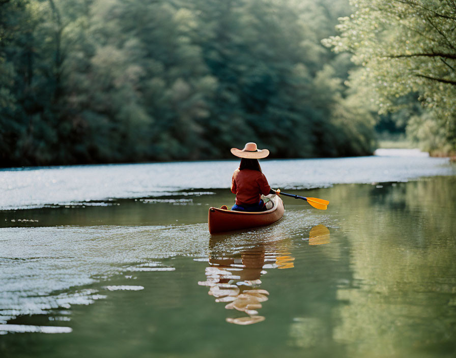 Person in wide-brimmed hat paddles red canoe on tranquil forest-lined river