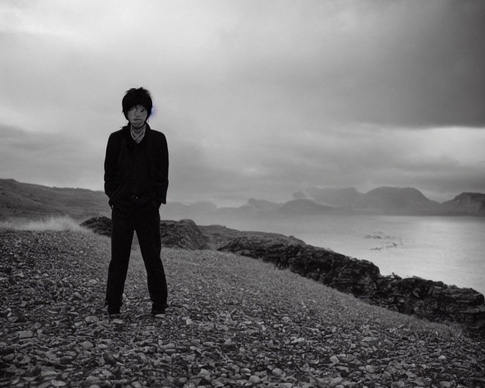 Person standing on pebbly shore with moody sea and distant mountains under overcast skies