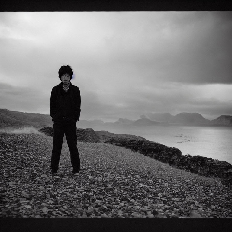 Person standing on pebbly shore with moody sea and distant mountains under overcast skies