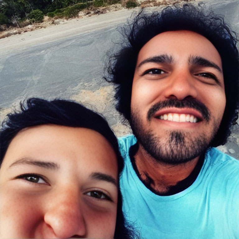 Two people smiling for a selfie outdoors under a clear sky with sandy path and greenery.