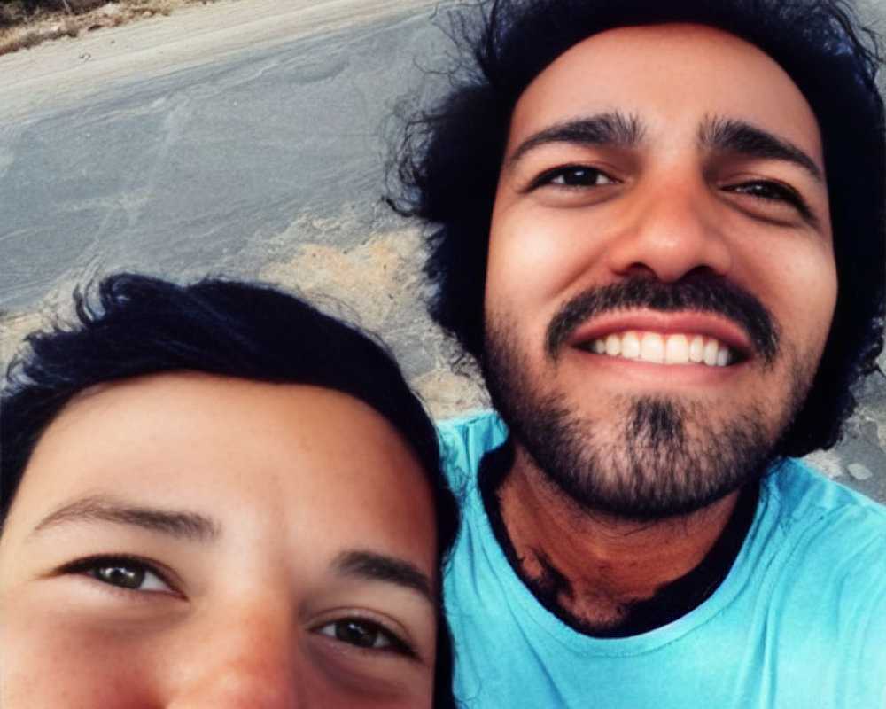 Two people smiling for a selfie outdoors under a clear sky with sandy path and greenery.