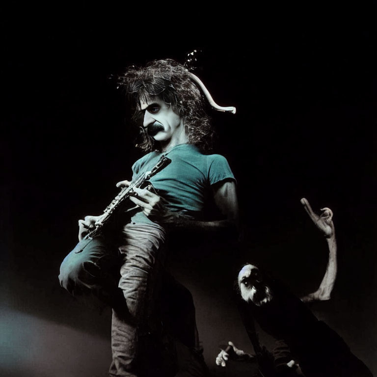 Mustached musician with curly hair plays guitar on stage with dramatic lighting, audience member's hand visible