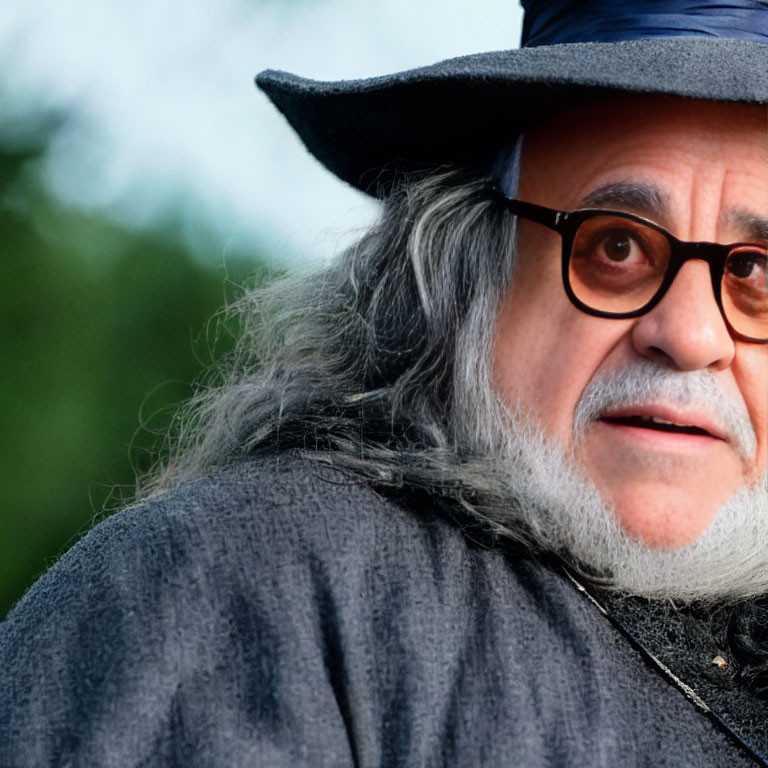 Gray-bearded man in glasses and hat gazes sideways with blurred background