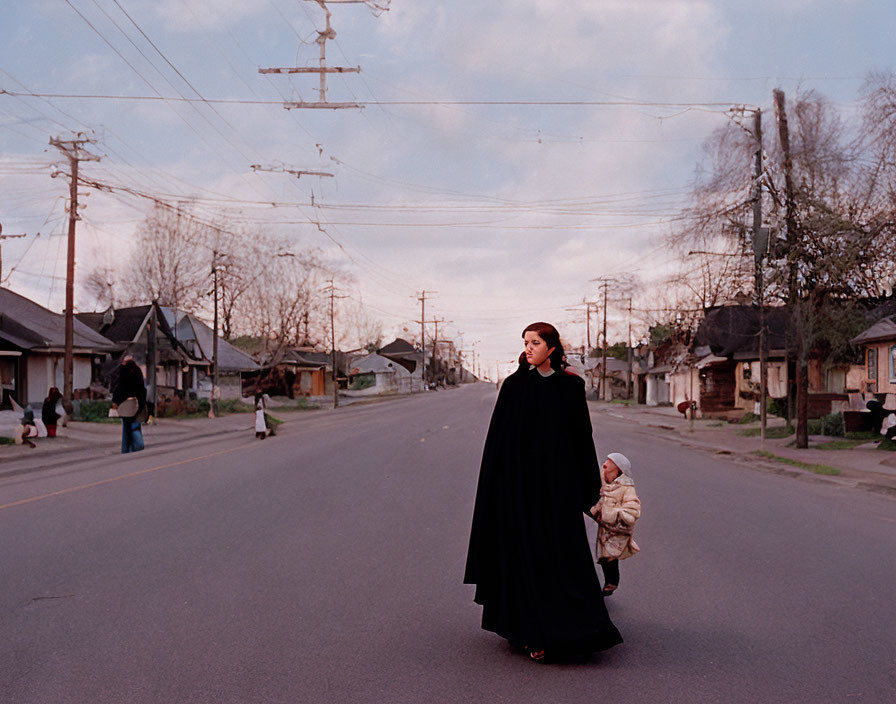 Woman in black cape walks with child on street at dusk.