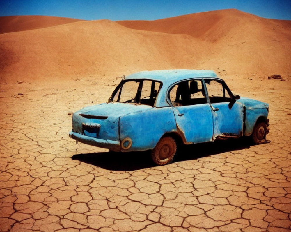 Abandoned blue car on cracked dry ground with sand dunes and clear sky