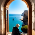 Elderly man squatting in sunlit archway overlooking serene blue sea.