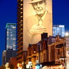 City street at dusk with large mural, vehicles in motion, and illuminated buildings.