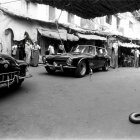 Monochrome image: cobra in street with vintage cars & people in market.