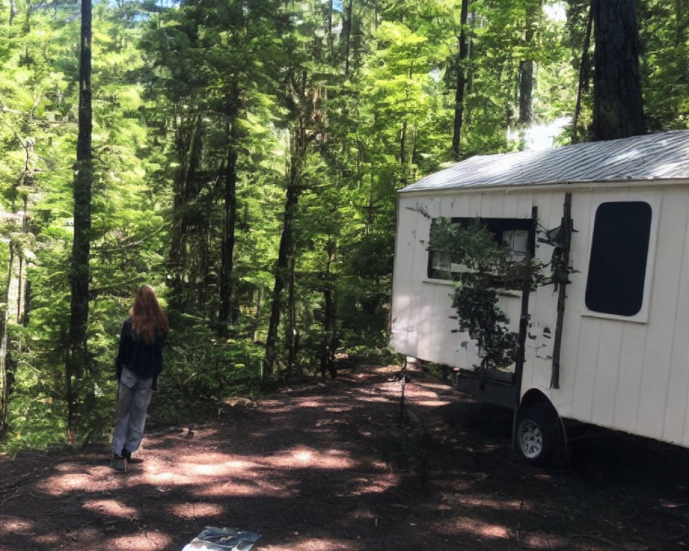 Long-Haired Person in Sunlit Forest Clearing Near White Trailer