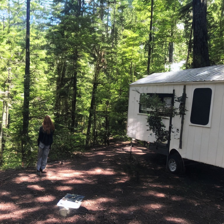 Long-Haired Person in Sunlit Forest Clearing Near White Trailer