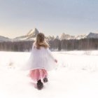 Girl in dress and hat walking in snowy field towards forest under soft glowing sky