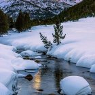 Snow-covered houses by river with warm glow lights, trees, and mountains at dusk