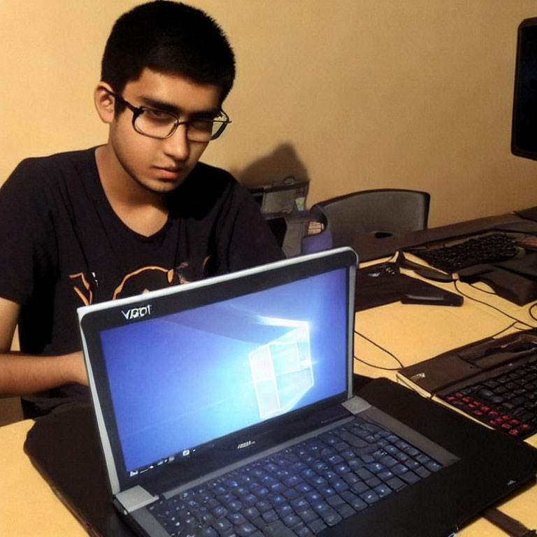 Young person with glasses at laptop and desktop computer in background.