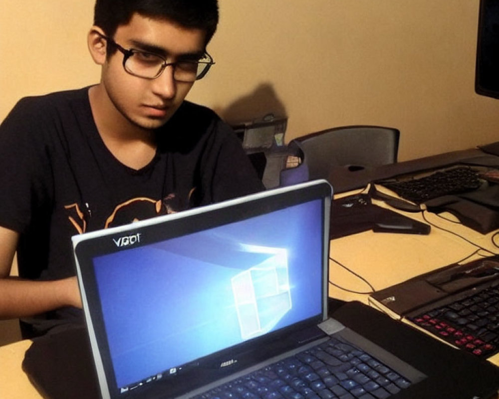 Young person with glasses at laptop and desktop computer in background.