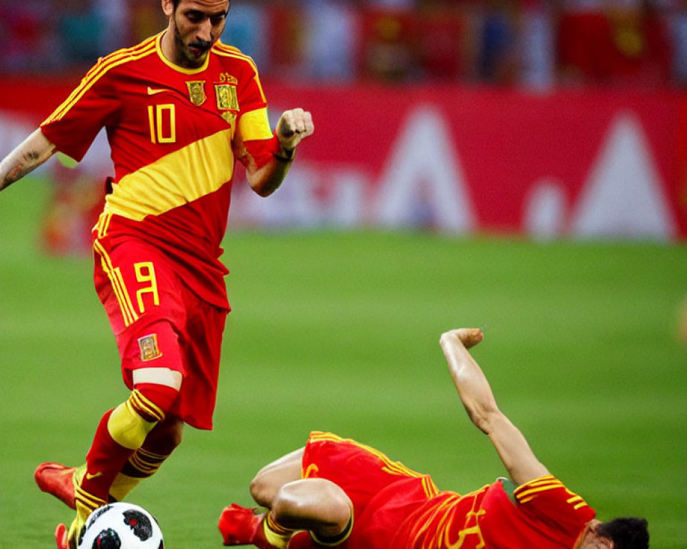 Soccer players in red and yellow kits on pitch: one standing, one fallen