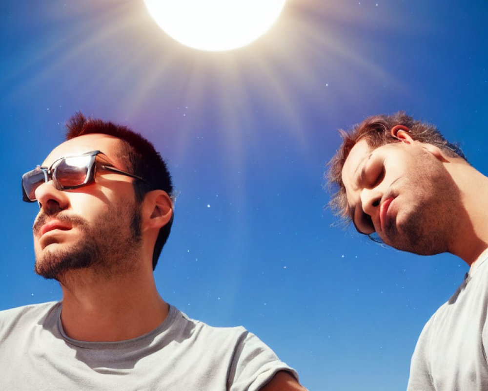 Two men with closed eyes in sunlight, one with sunglasses, under blue sky