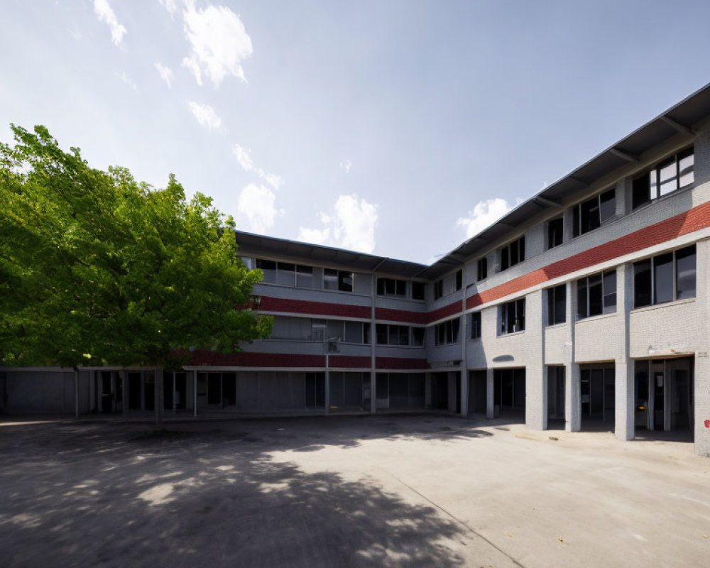 Two-story U-shaped building in empty school courtyard under blue sky with large green tree.