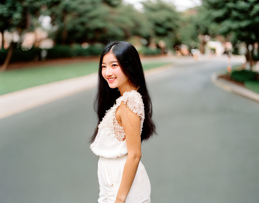 Smiling young woman with long dark hair in white top on tree-lined road