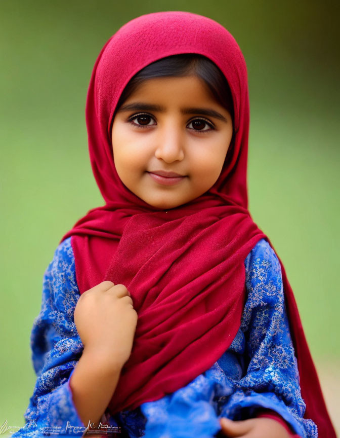 Smiling young girl in red hijab and blue dress on green background