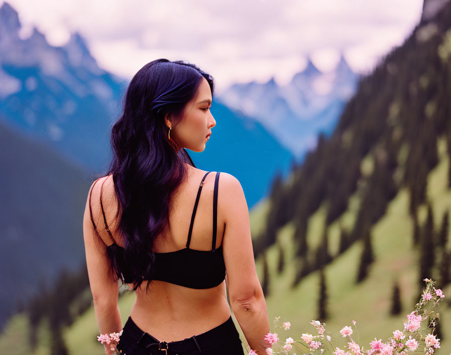 Woman in black top surrounded by wildflowers and mountains, gazing contemplatively.