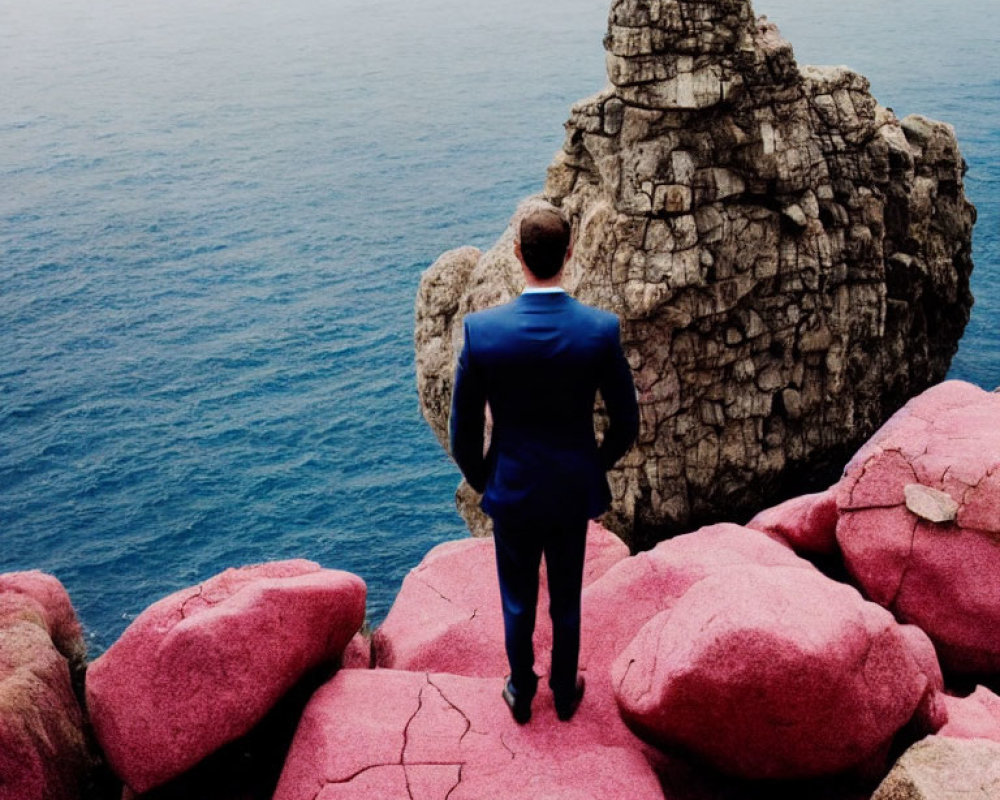 Man in Blue Suit on Pink Rocks Gazes at Distant Figure on Tall Rock by the Sea