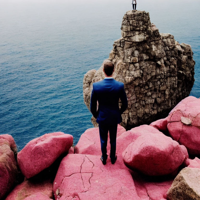 Man in Blue Suit on Pink Rocks Gazes at Distant Figure on Tall Rock by the Sea