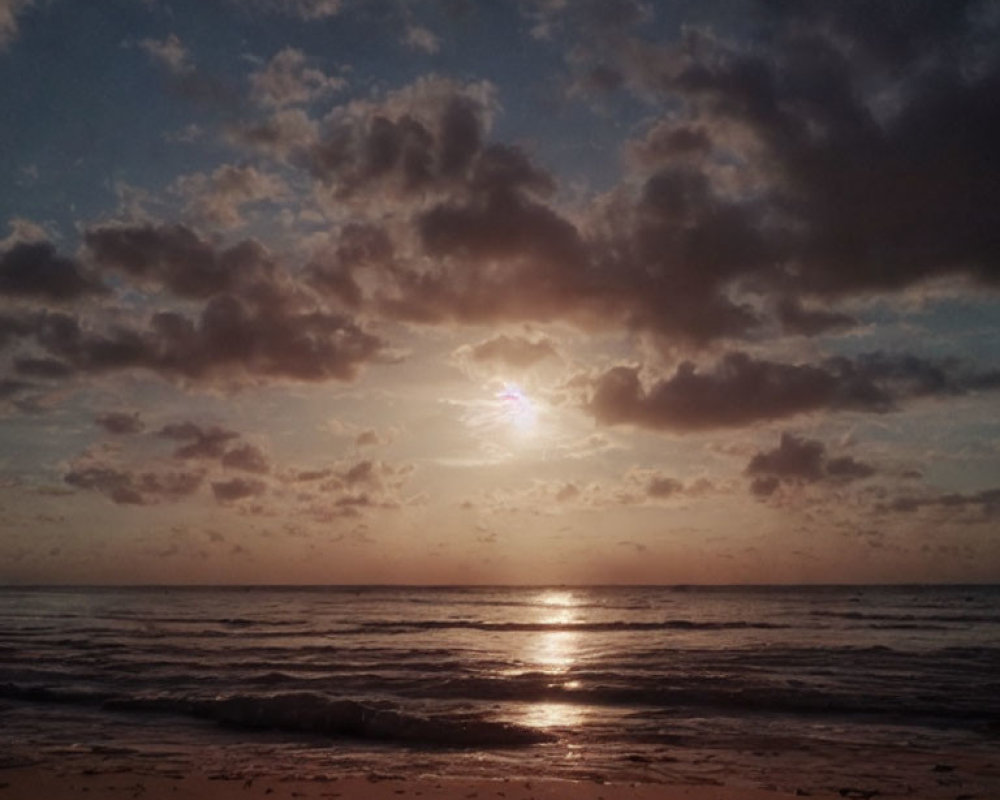 Ocean sunset with sun obscured by clouds reflecting on water and beach.