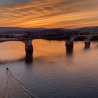 Sunset painting of bridge over water with orange skies and silhouetted buildings